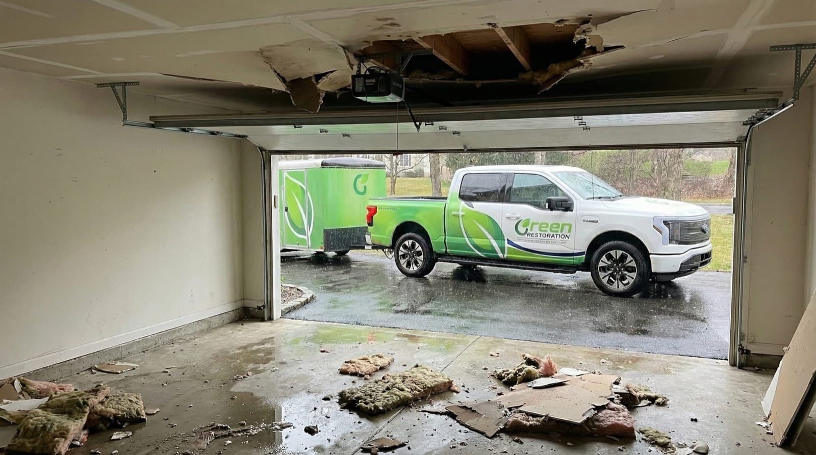 Collapsed water-damaged garage ceiling with fallen insulation and drywall debris, a branded Green Restoration pickup truck and response trailer parked outside